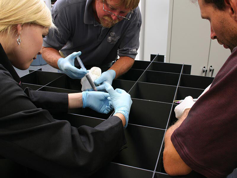 Near zero CTE antenna segment being manufactured. Image shows a team of three people working on a large carbon fiber grid and applying adhesive to a corner connection.